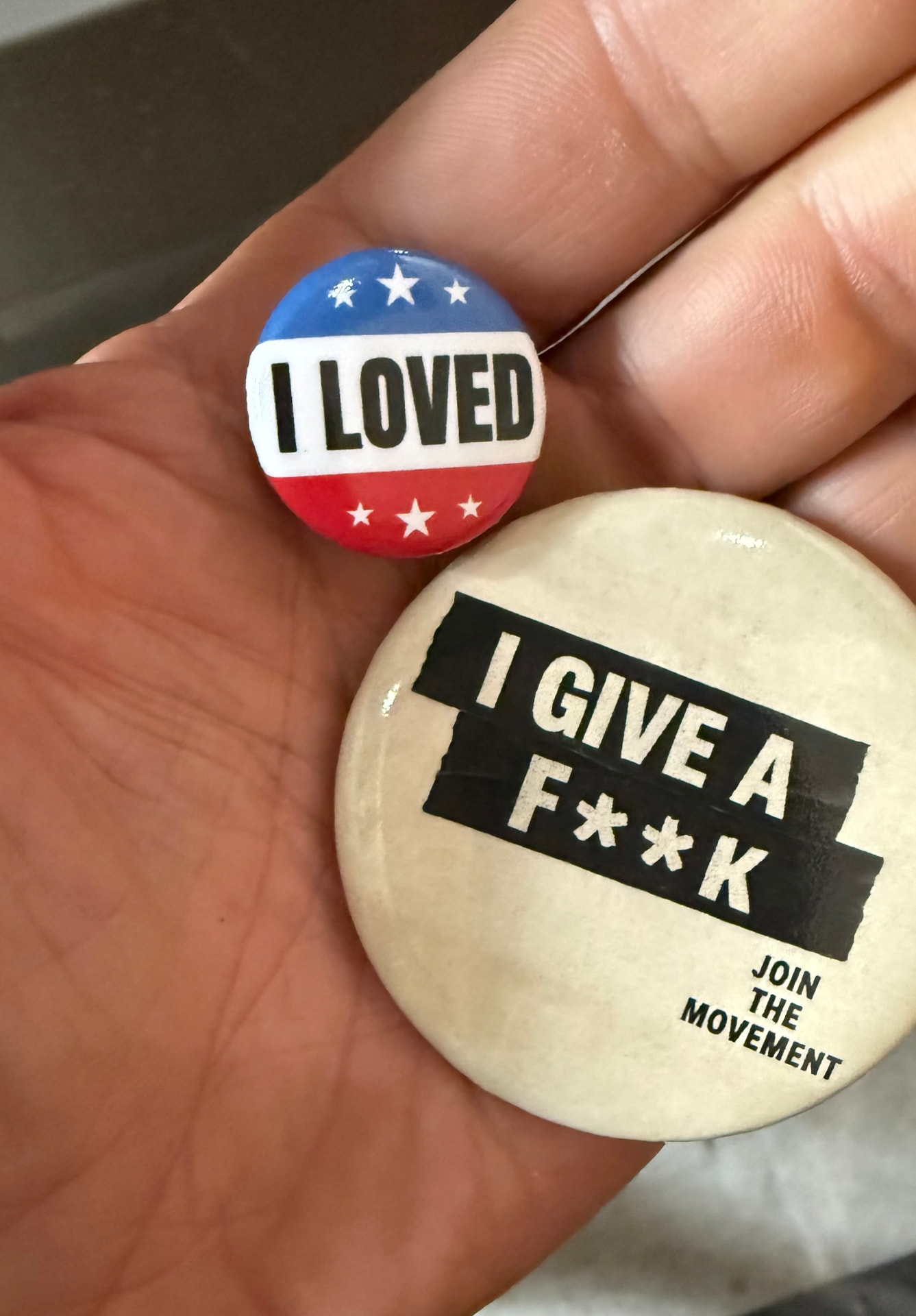 Both pins held in an open hand: the small red, white and blue I LOVED voter-sticker pin and the larger off-white I GIVE A F**K button with black tape lettering